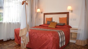 Een netjes opgemaakt bed met een rode sprei en meerdere kussens, een nachtkastje met een lamp, en dunne gordijnen voor de ramen.