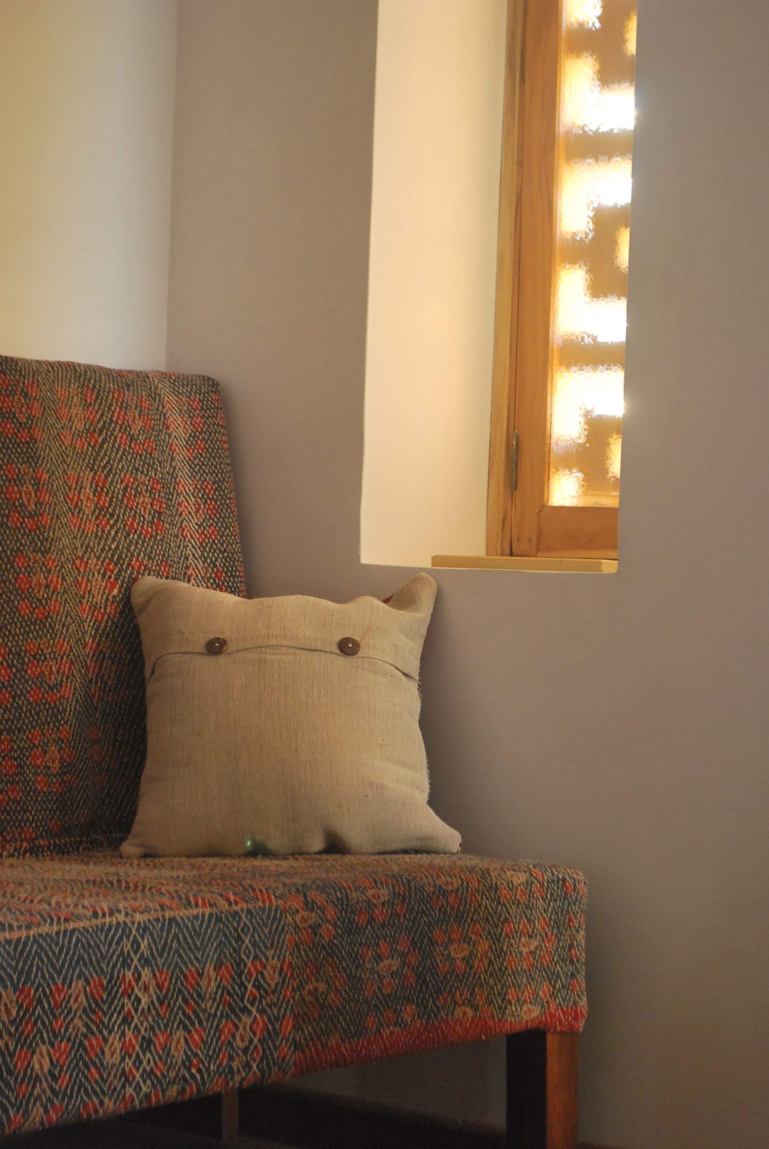 Un rincón de una habitación con un sillón estampado y un cojín, una ventana con marcos de madera y una pared lisa.