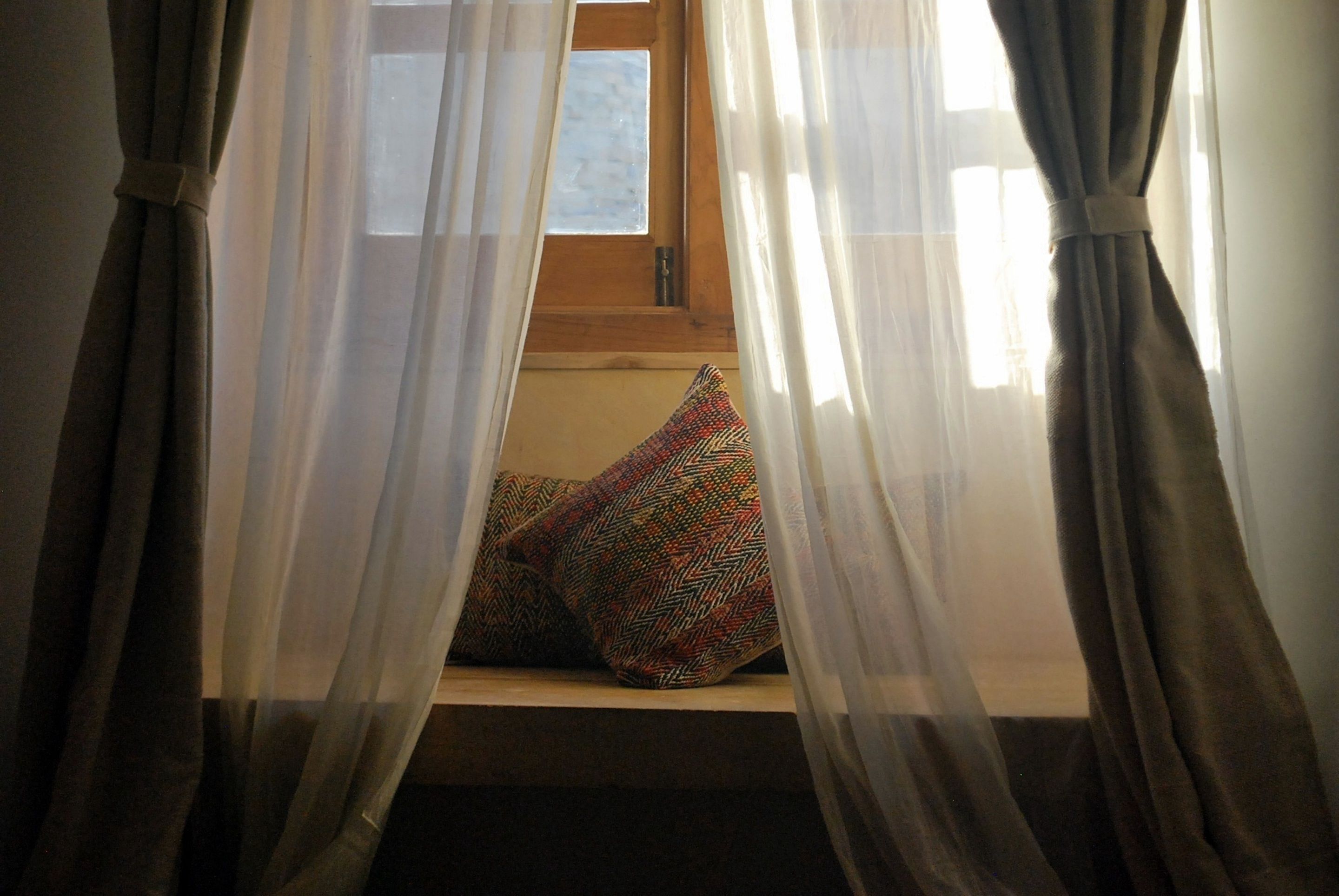 A window with sheer curtains, a wooden frame, and a colorful cushion on a ledge.