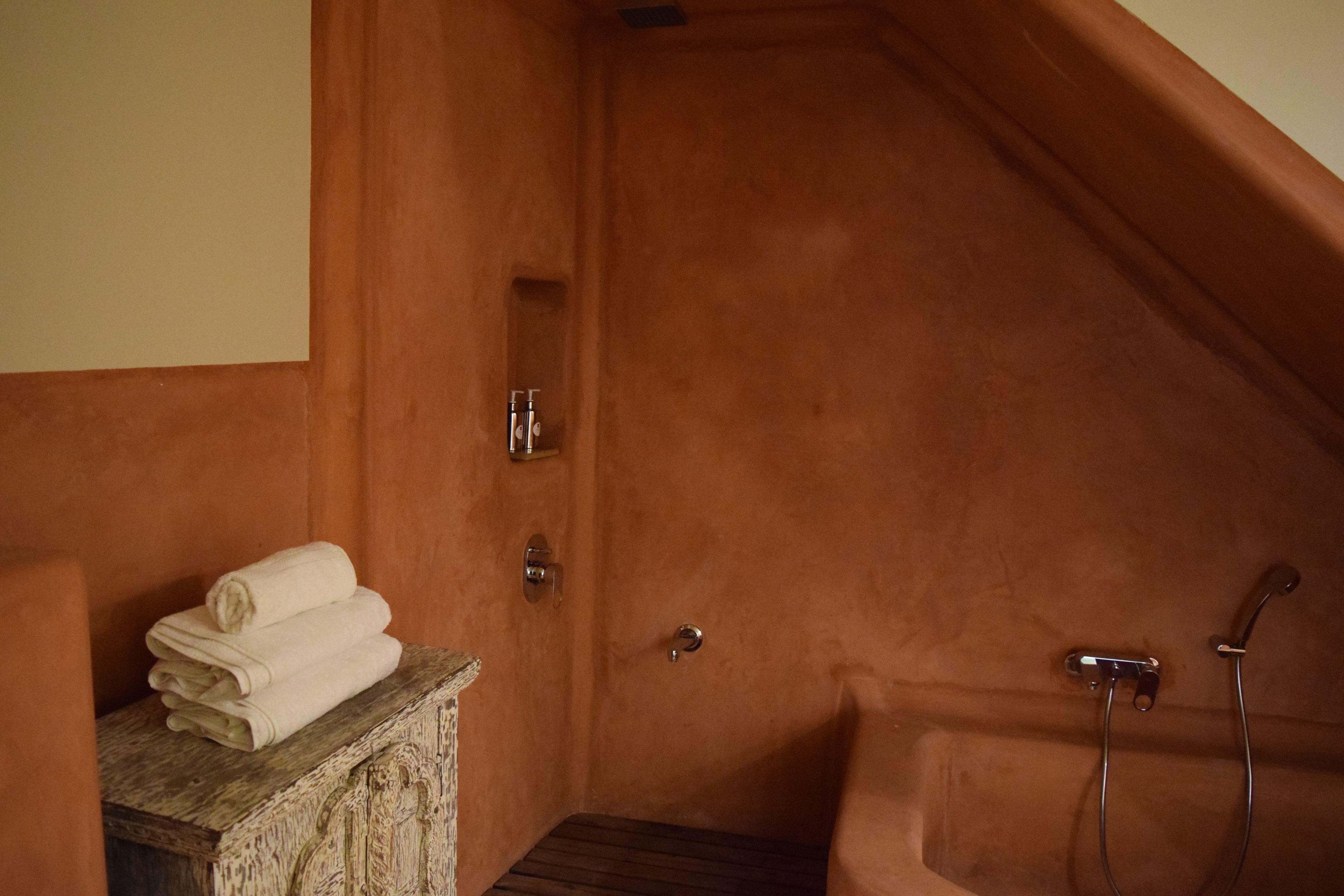 A small, enclosed bathroom with a wooden bench, a stack of towels, and a bathtub with a shower head.