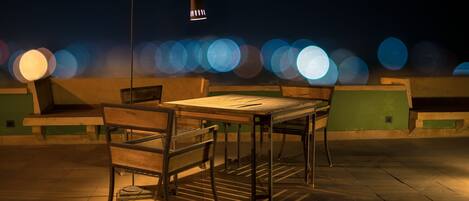 An outdoor seating area with a table and chairs, a lamp, and a view of blurred lights in the background.