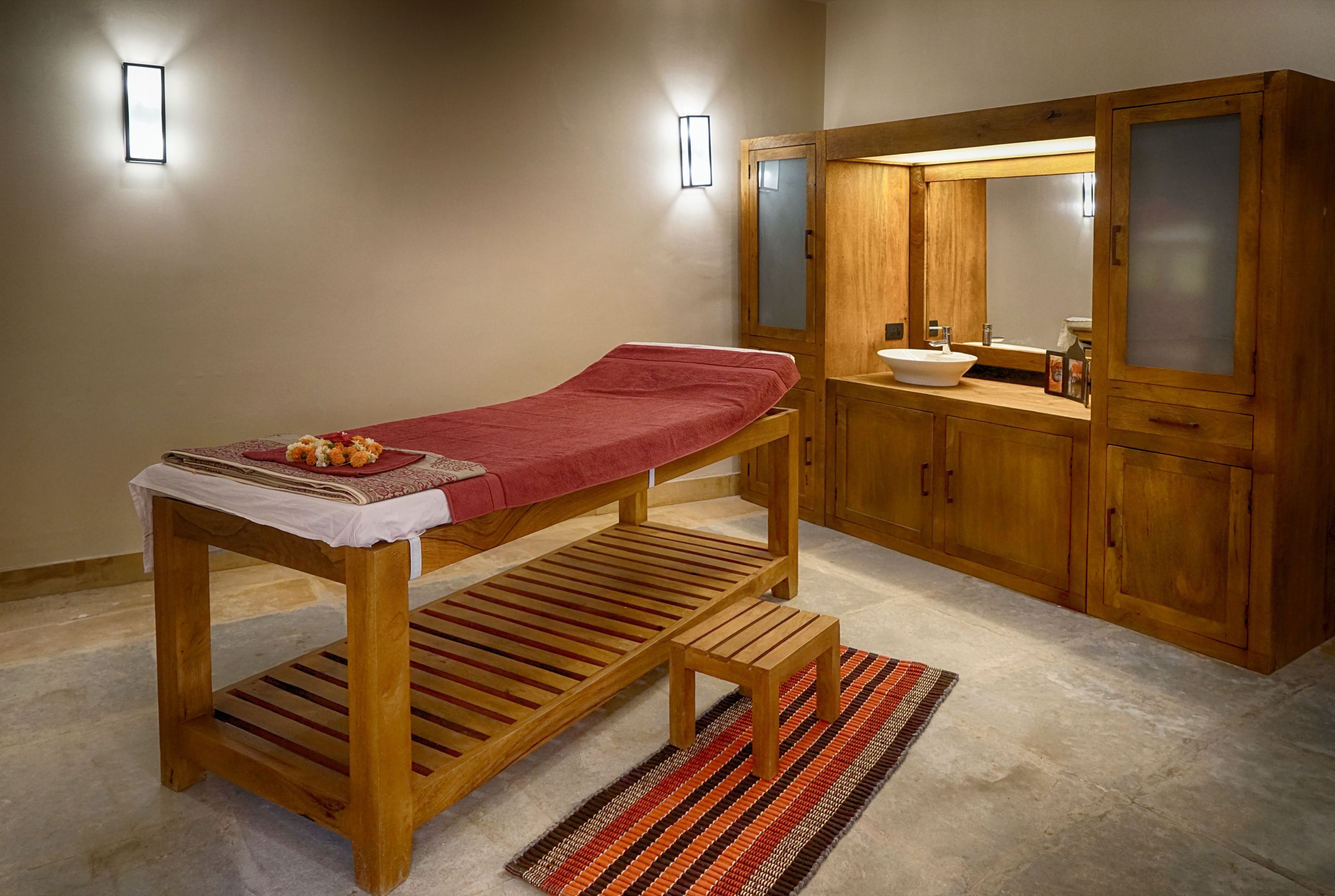 A wooden massage table with a red towel, a small wooden stool, and a sink with a mirror in a wooden cabinet.