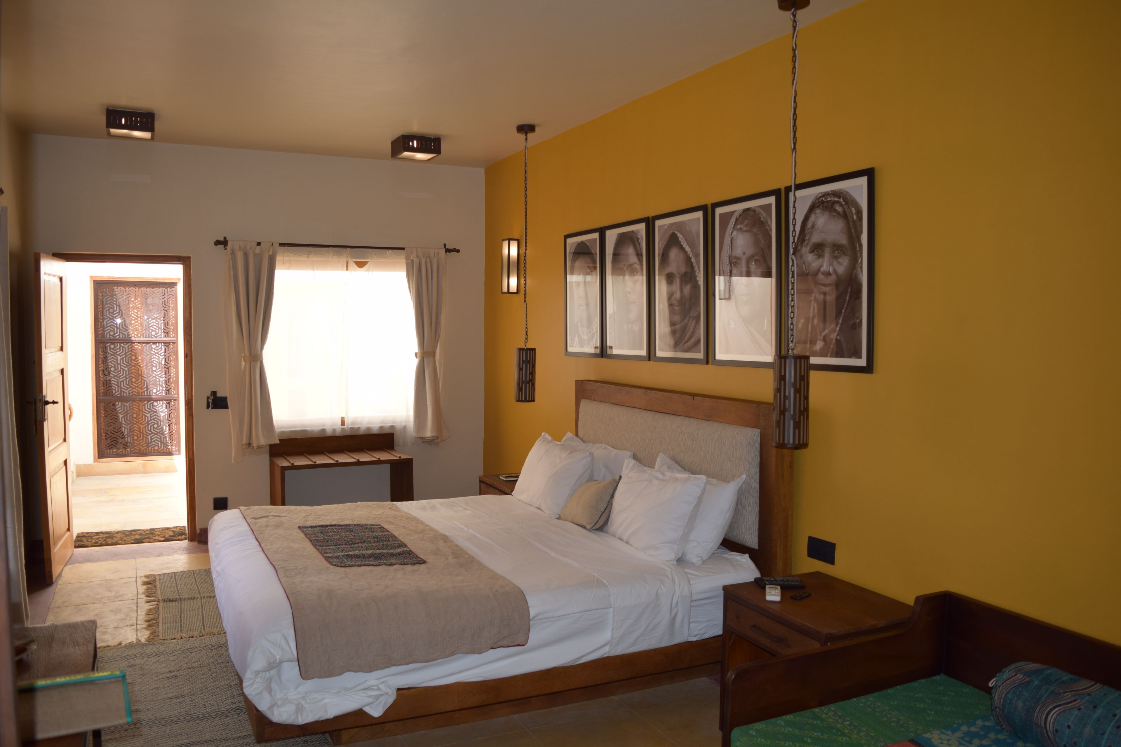 A bedroom with a bed, bedside tables, a nightstand, a window with curtains, and a door leading to another room.