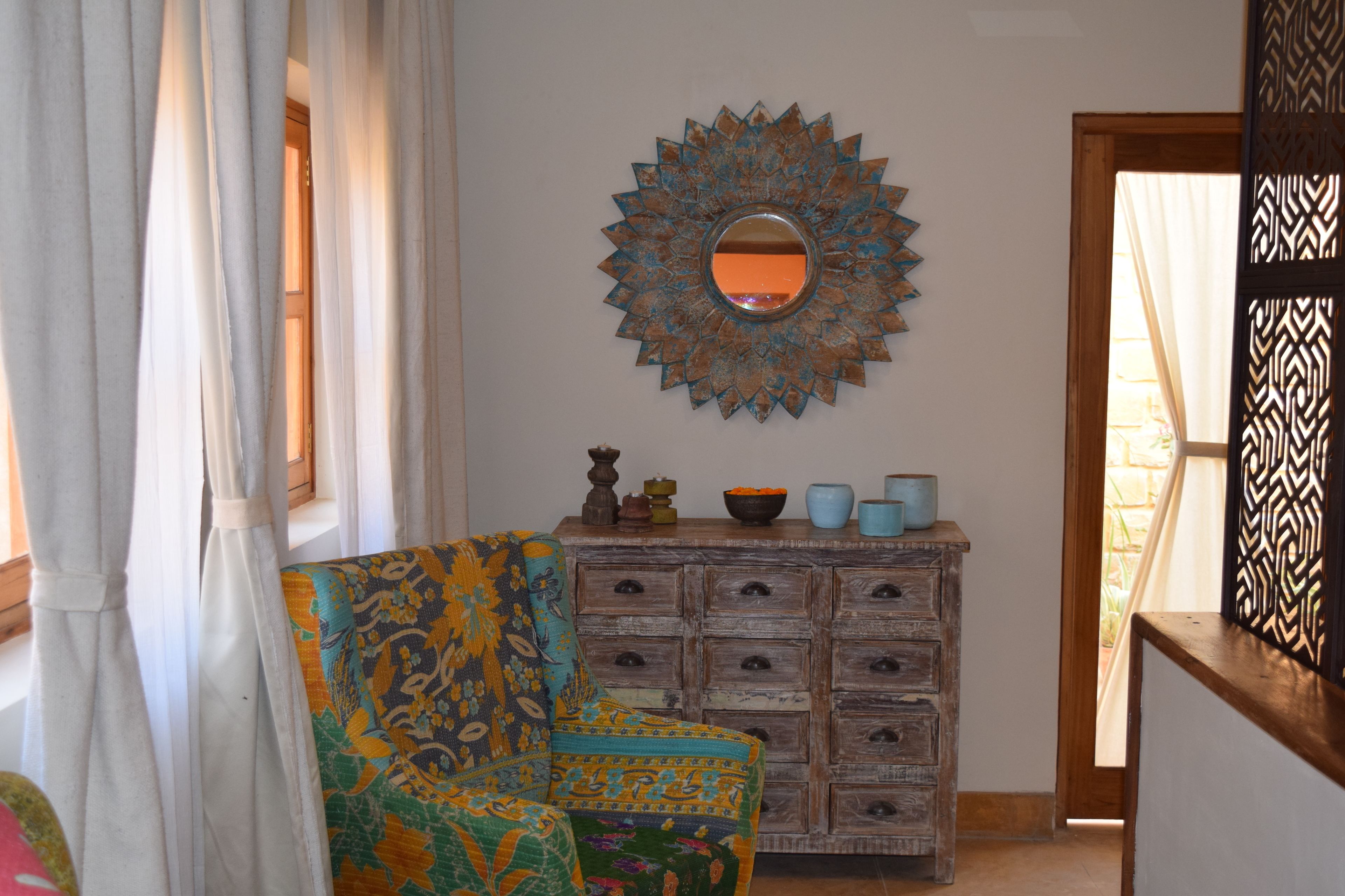 A room with a patterned armchair, a wooden dresser with drawers, and a decorative mirror on the wall.