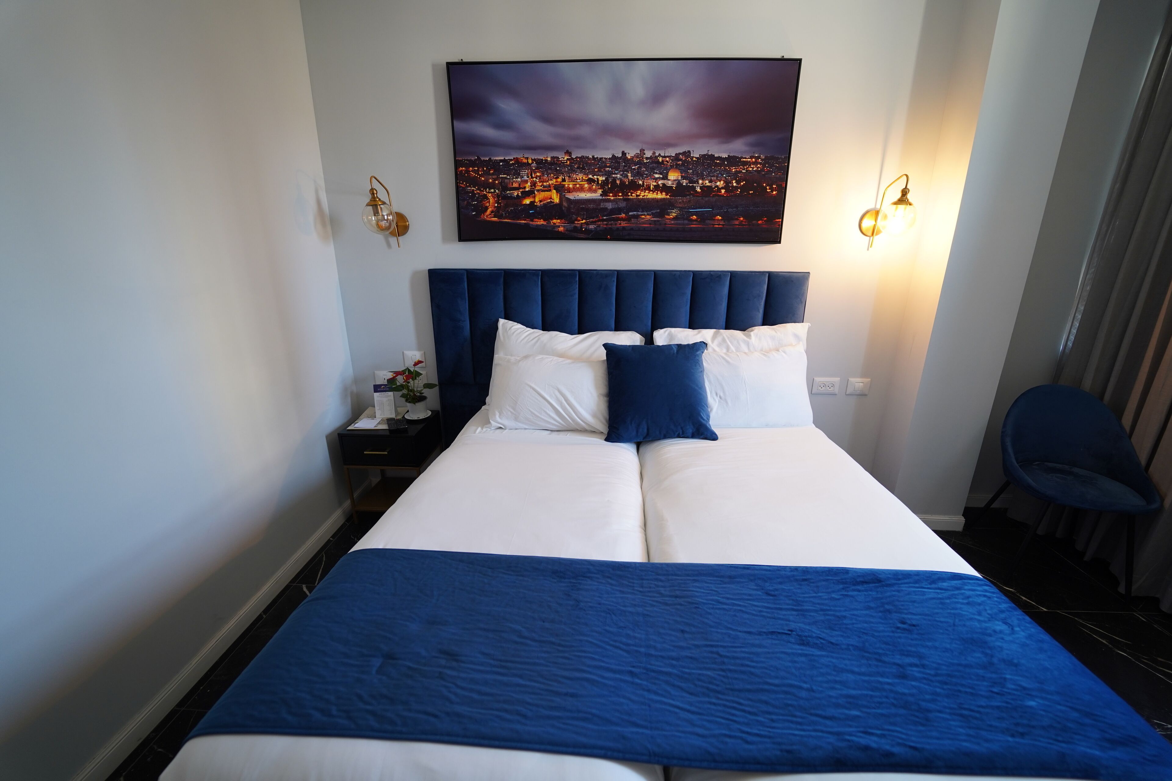 Habitación de hotel con una cama grande, un cuadro de paisaje urbano sobre la cama, dos lámparas de noche y una silla.