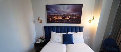 Habitación de hotel con una cama grande, un cuadro de paisaje urbano sobre la cama, dos lámparas de noche y una silla.
