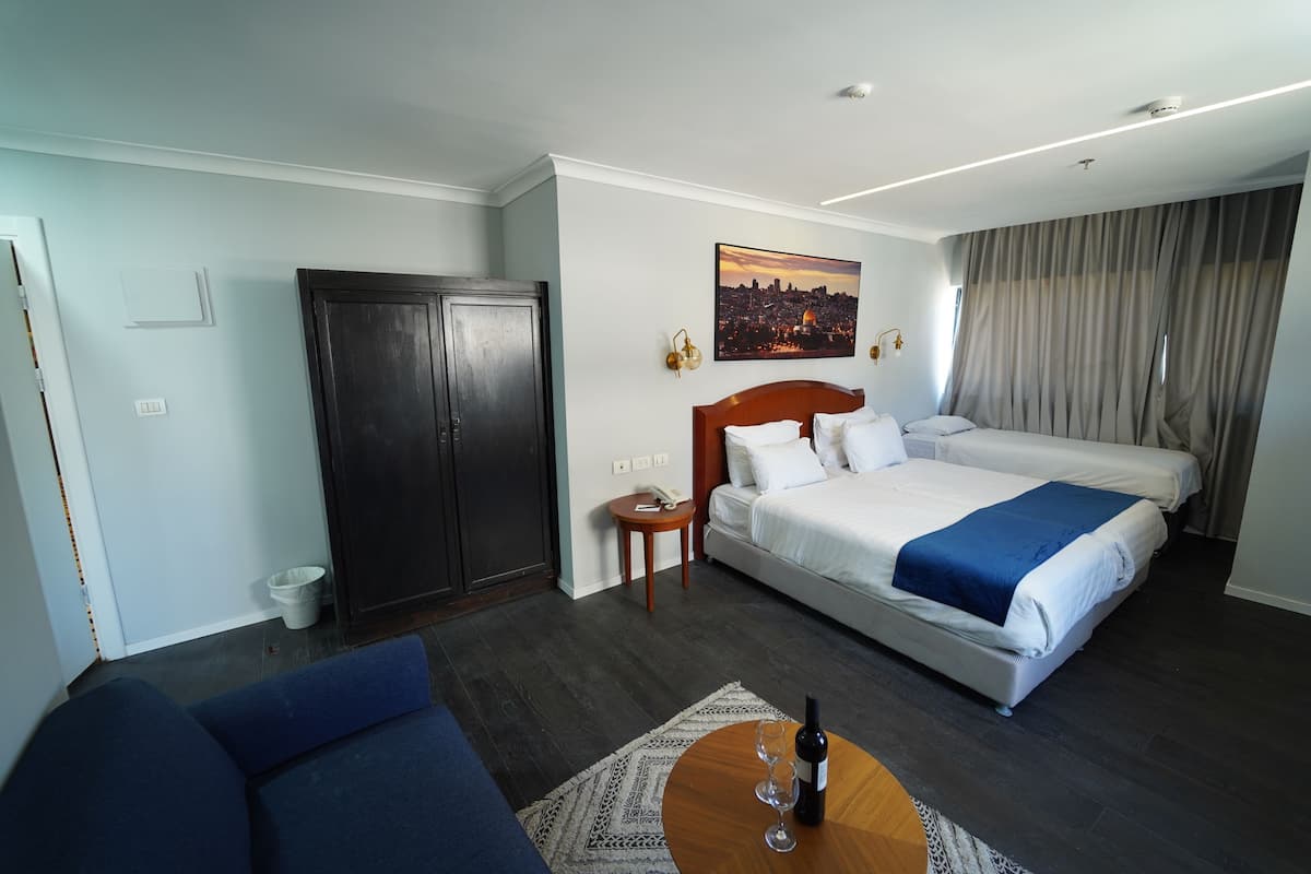 Une chambre d’hôtel avec un lit, un canapé, une table en bois sur laquelle se trouvent une bouteille et des verres, une armoire, un tableau représentant un paysage urbain et une vue sur la ville par la fenêtre.