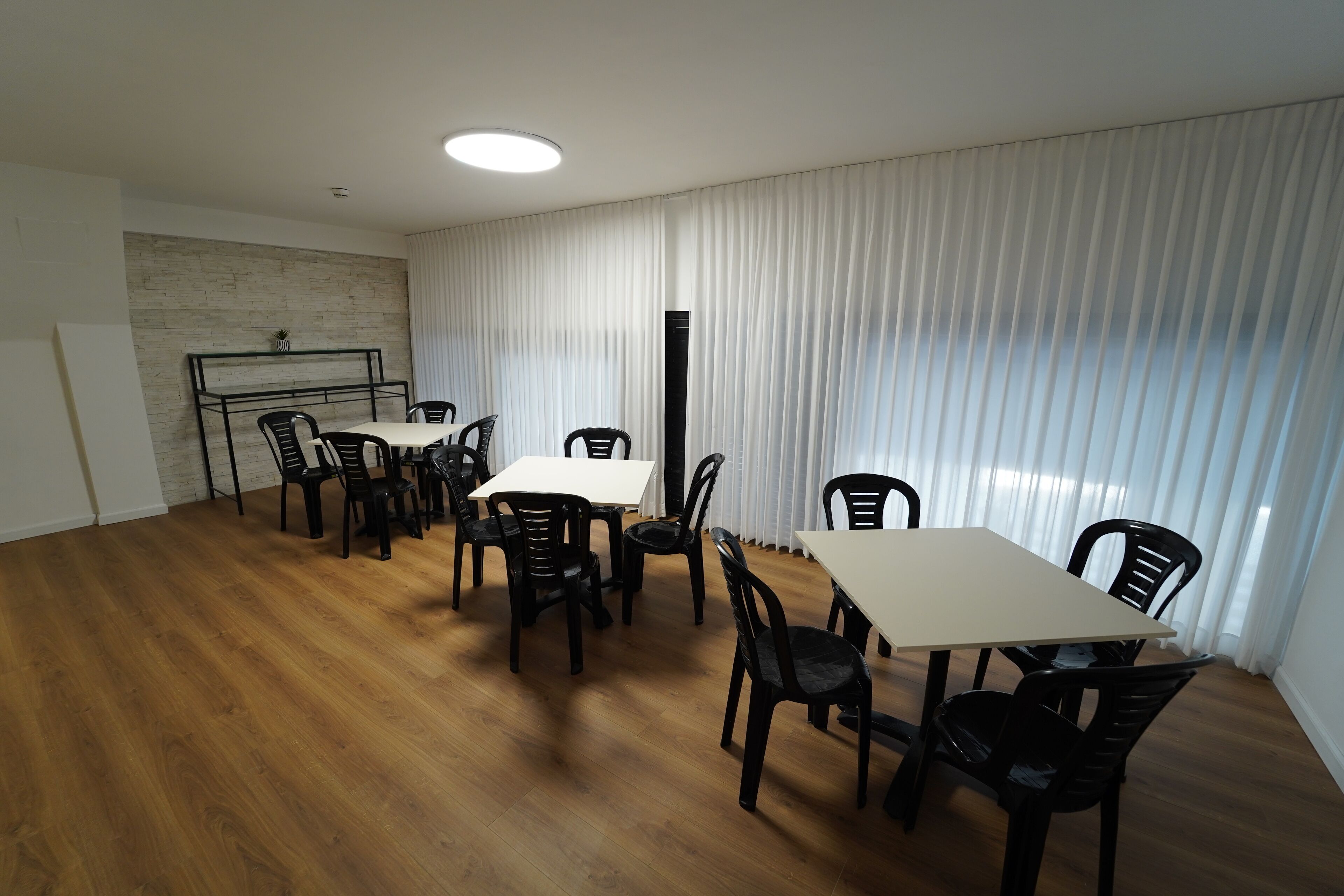 Zona de dinar amb sòl de fusta, parets blanques i una paret decorativa de maó. Hi ha dues taules de dinar amb cadires, i un finestra gran amb cortines blanques.