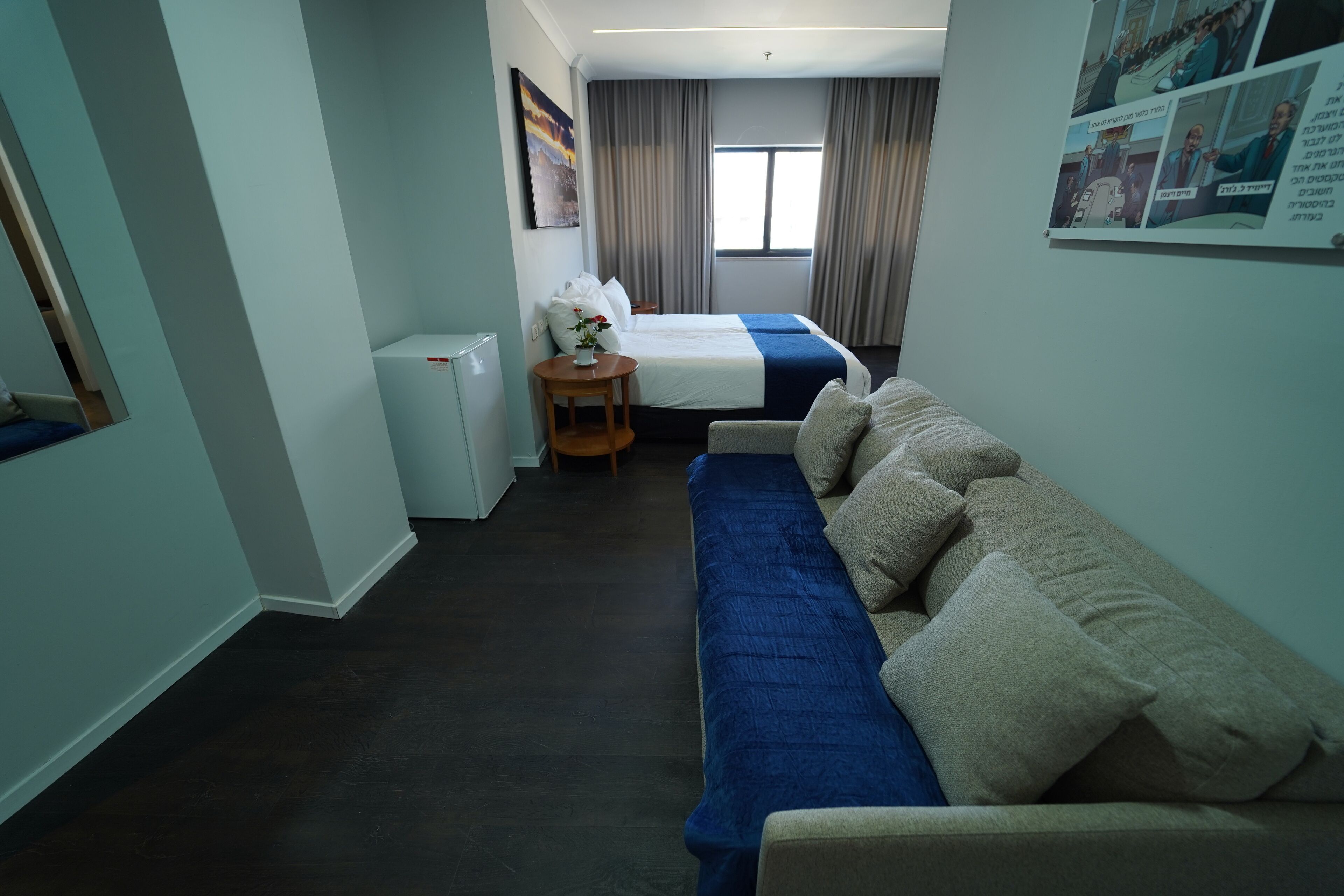 A hotel room with a sofa, a bed, a small table with a vase, and a refrigerator.