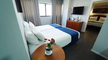 A hotel room with a bed, a small table with a plant, a TV on the wall, and a painting above the bed.