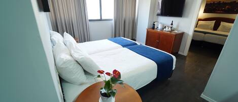Habitación de hotel con una cama, una mesita con una planta, un televisor en la pared y un cuadro sobre la cama.