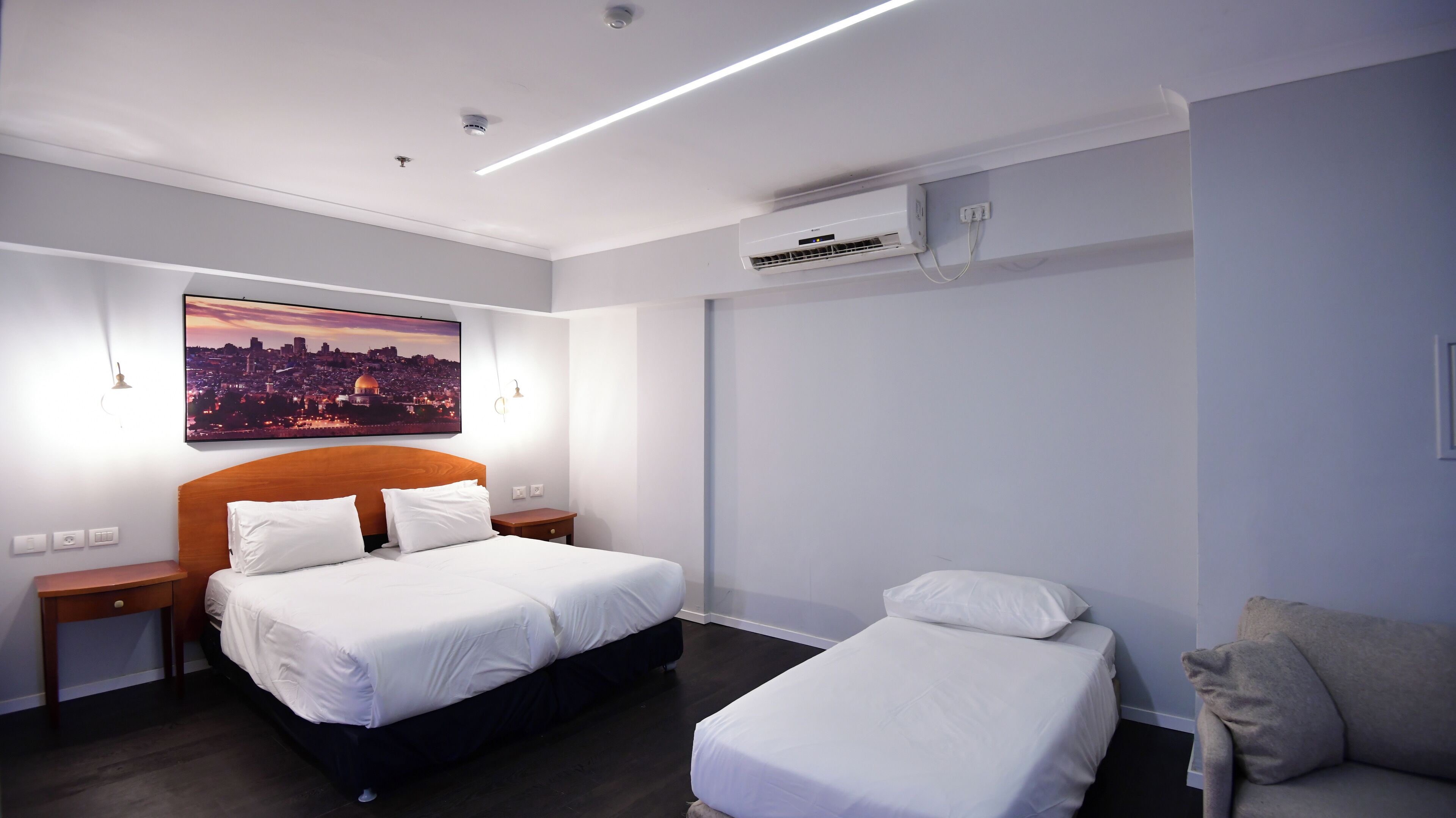 Ett hotellrum med en stor säng, en mindre säng, en soffa, en luftkonditioneringsanläggning, ett nattduksbord, en lampa och en tavla med en stadsbild på väggen.