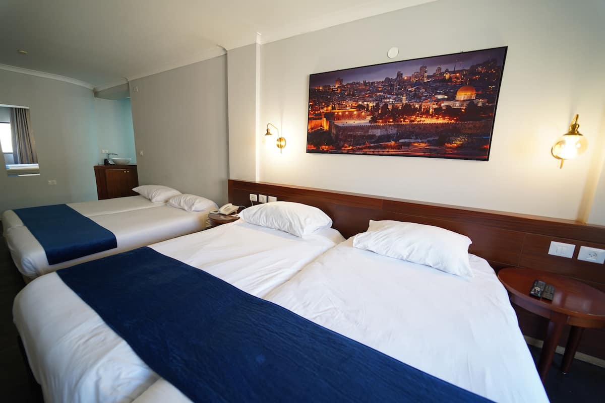 Une chambre d’hôtel avec deux lits, une table de chevet, une lampe fixée au mur et un tableau représentant un paysage urbain.