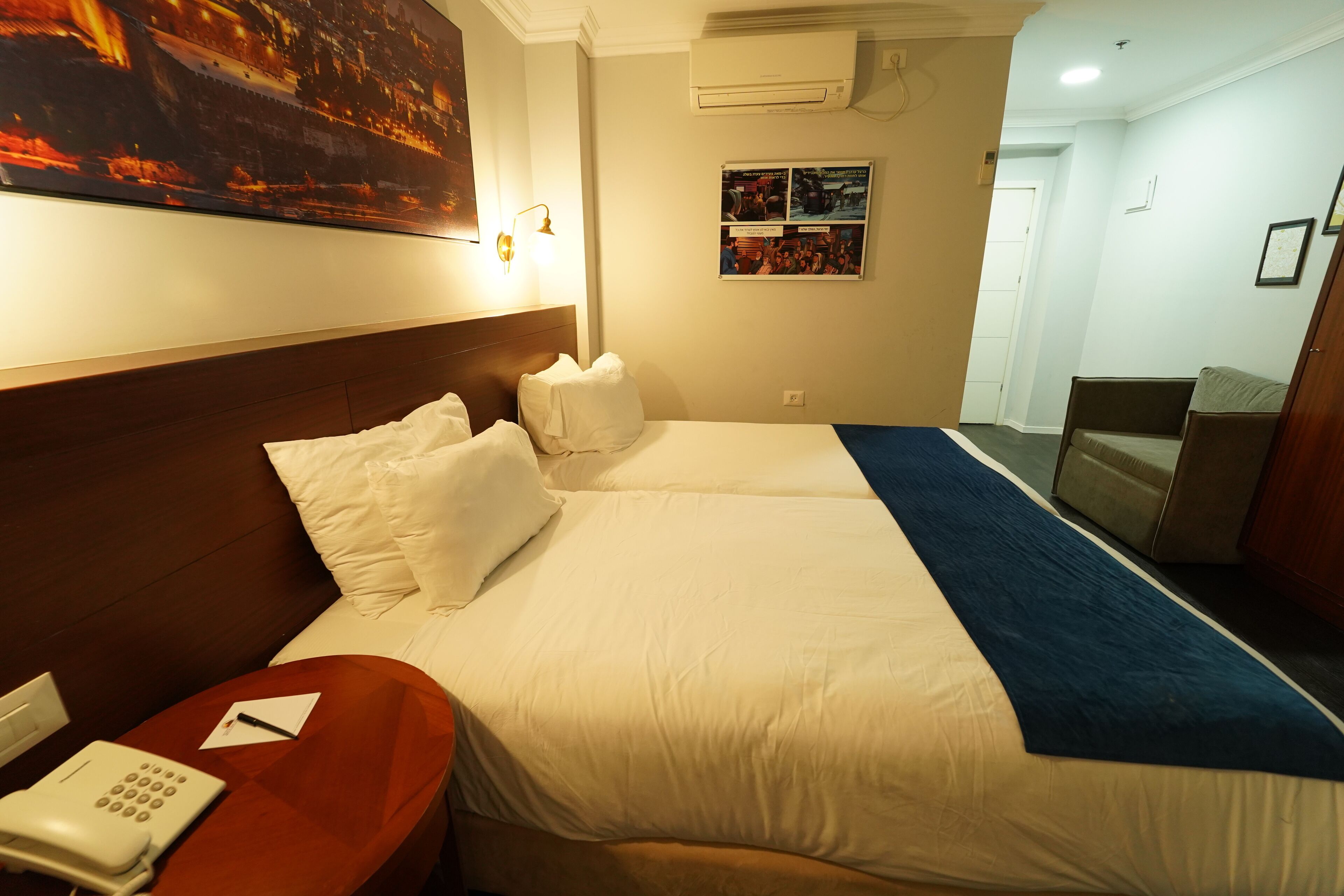 Una habitación de hotel con una cama grande, una mesita de noche con teléfono, aire acondicionado en la pared y un cartel en la pared.