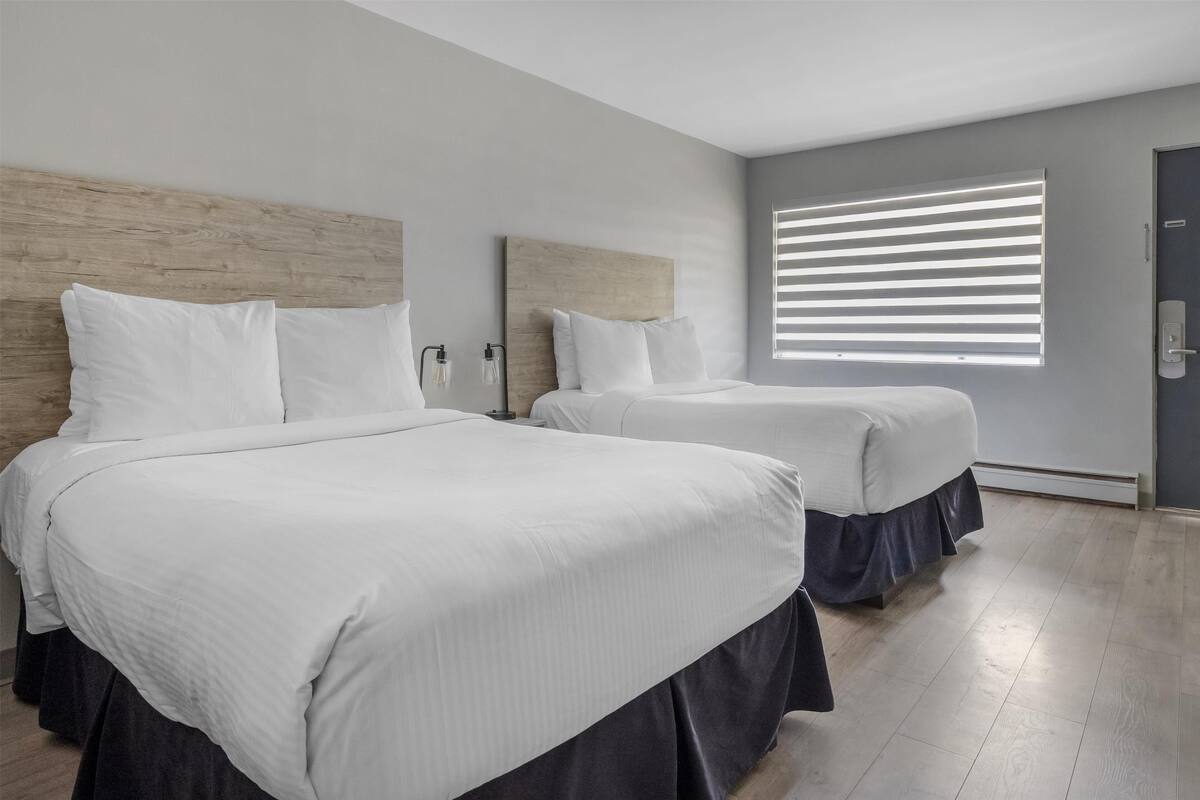 A modern hotel room with two beds, a large window with blinds, and a wooden headboard.