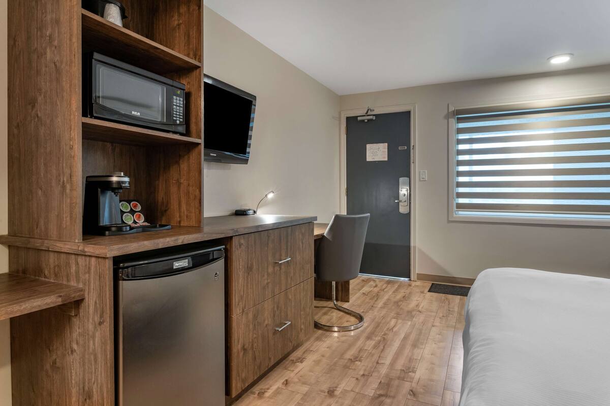 A compact hotel room with a wooden kitchenette, a flat-screen TV, a small desk with a chair, and a bed.