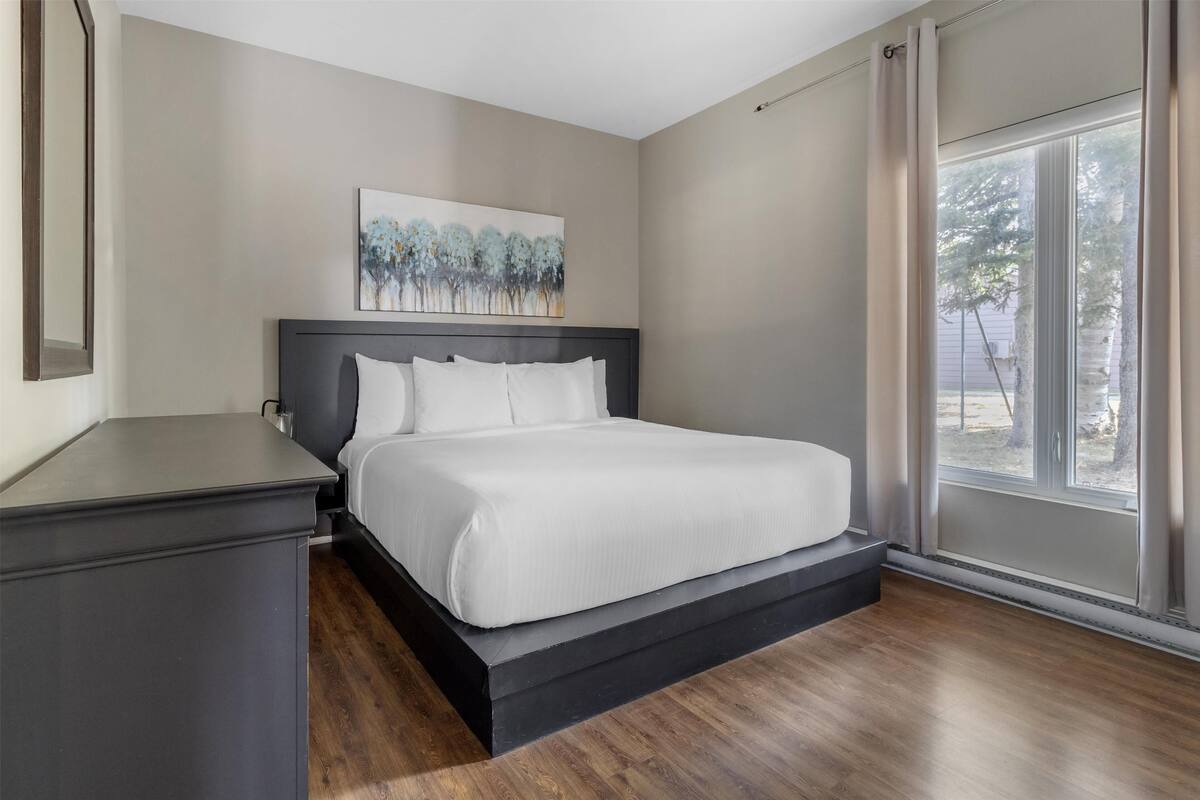 A bedroom with a bed, a nightstand, a painting on the wall, and a window with curtains.
