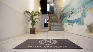 Un hall d’hôtel avec un sol en marbre, un escalier, une grande plante en pot et une fresque représentant un bateau sur le mur.