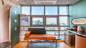 A modern room with a sofa, a small table, a kitchenette, and large windows offering a city view. - L&L Central Hotel (Jeju City)