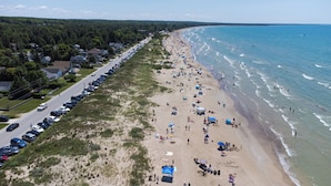 Une plage bordée d’une rangée de voitures garées le long de la route, une plage de sable où se trouvent des gens et des parasols, et une vue dégagée sur l’océan.