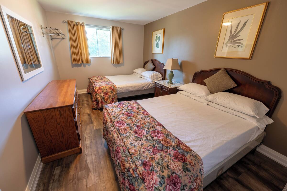 Ein Hotelzimmer mit zwei Betten, einem Holzkleiderschrank, einem Nachttisch mit Lampe und einem Fenster mit Vorhängen.