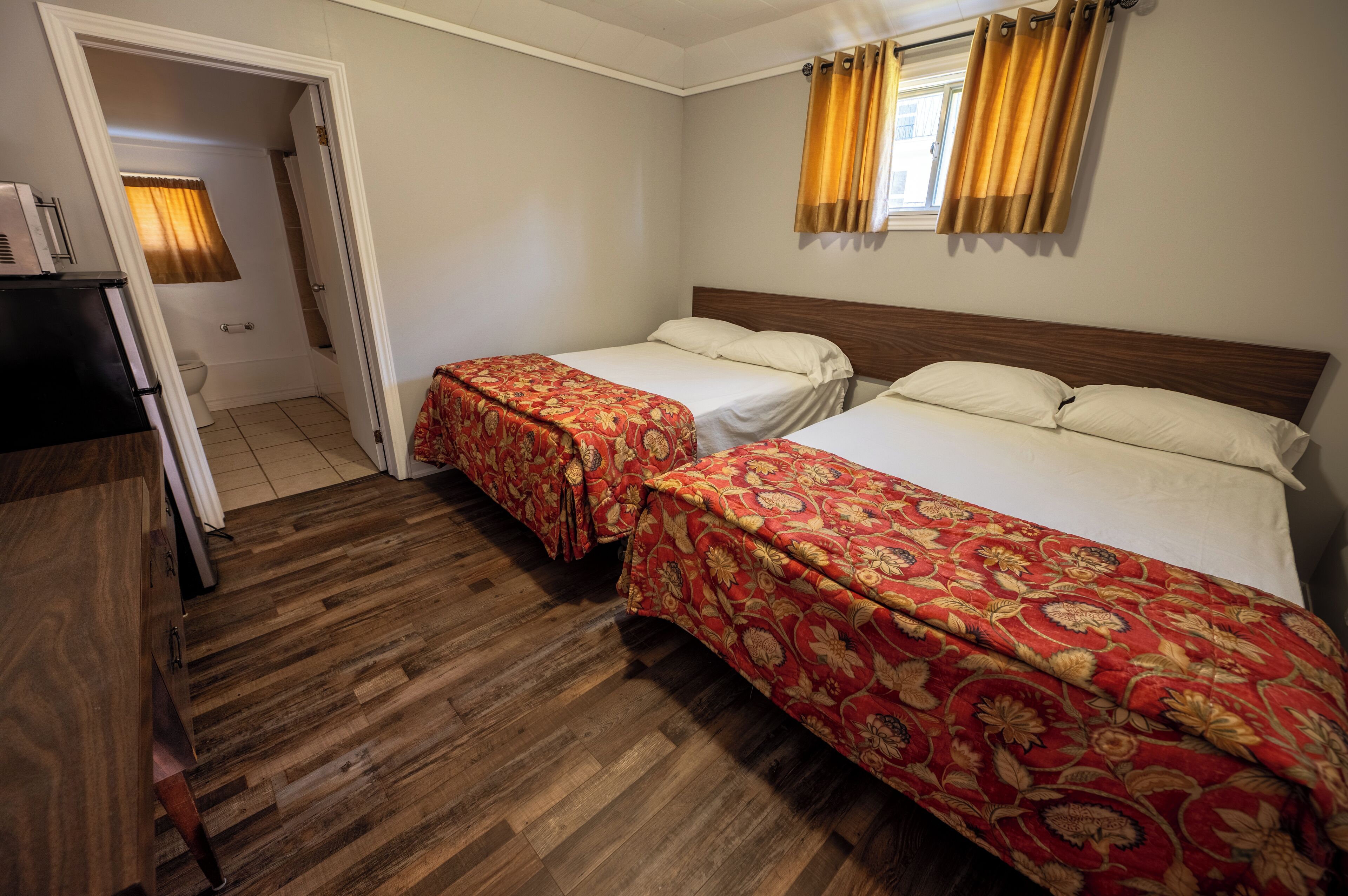 A hotel room with two beds, wooden flooring, a wardrobe, and a bathroom visible through an open door.