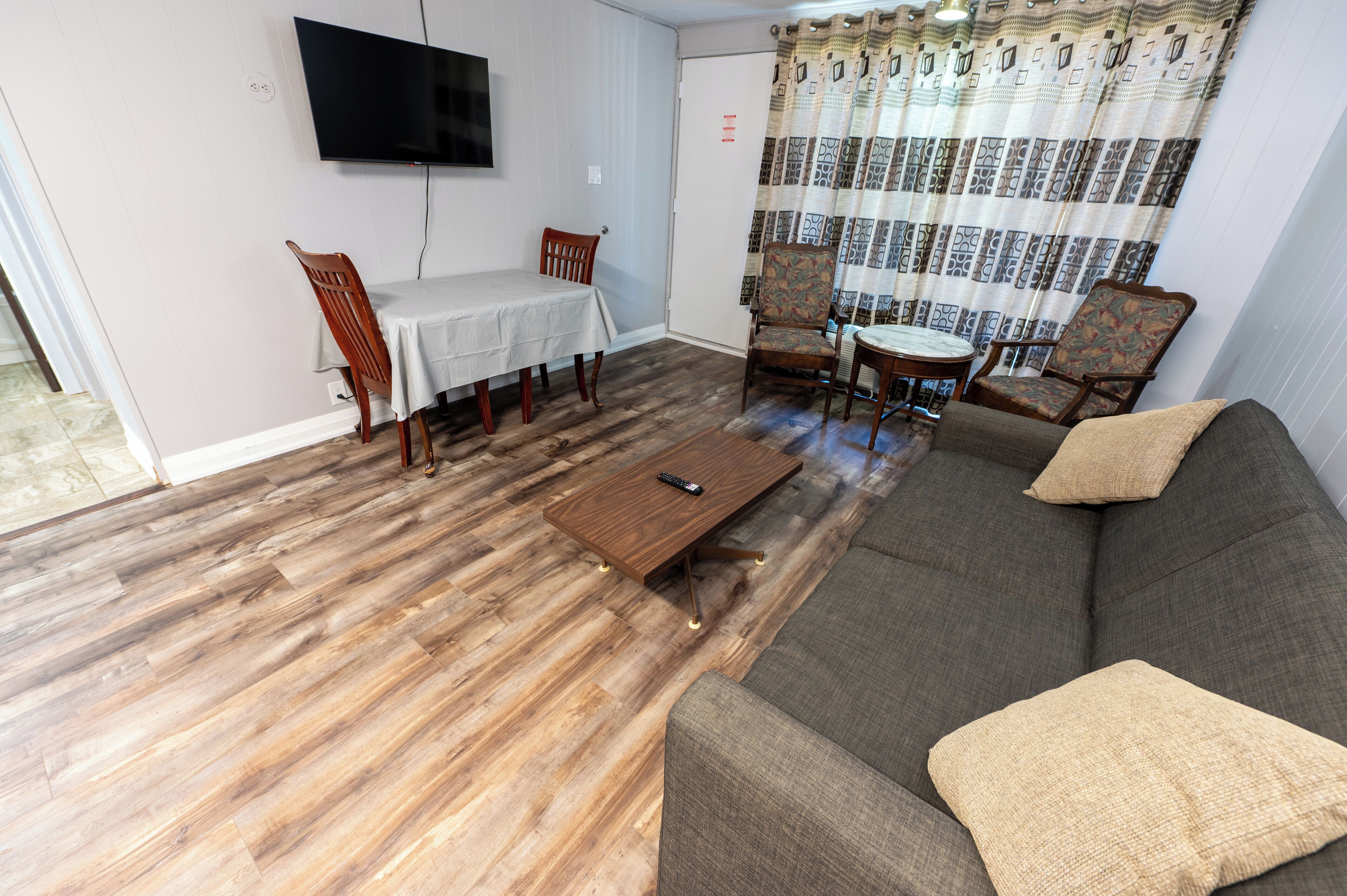 A living room with a wooden floor, a grey sofa, a coffee table, a TV mounted on the wall, and a dining table with chairs.