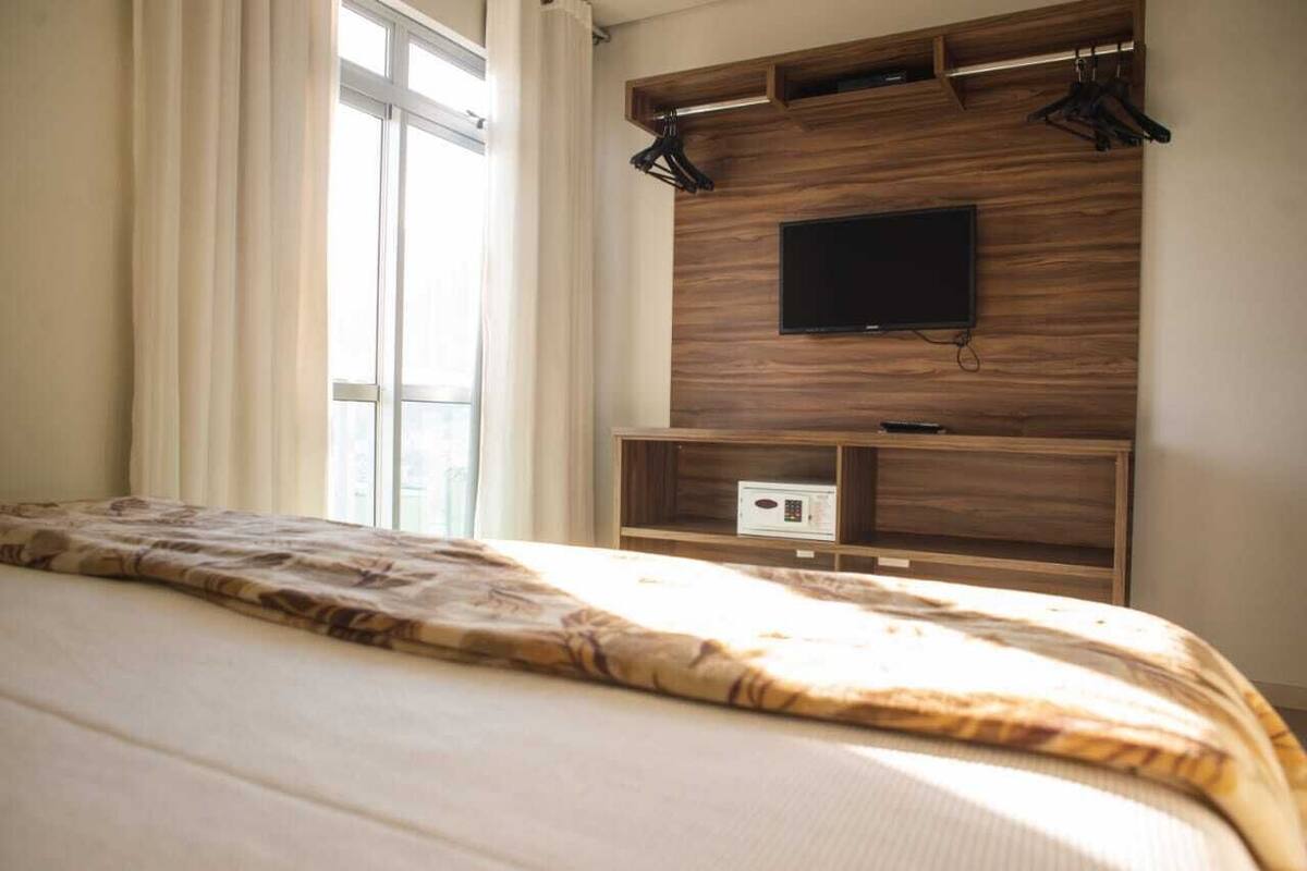 A hotel room with a bed, a TV mounted on a wooden wall, and a window with curtains.