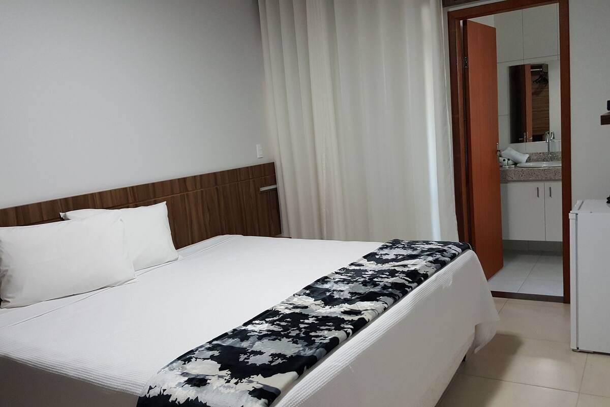 A hotel room with a bed, a wooden headboard, a white curtain, a bathroom with a sink and mirror, and a refrigerator.