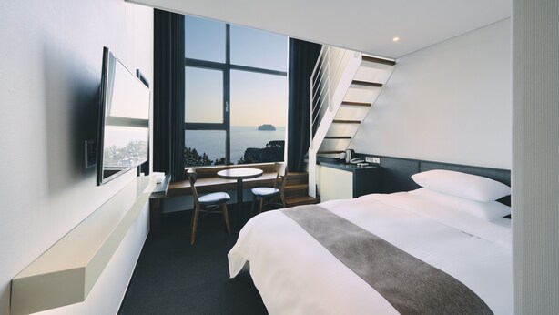 A modern hotel room with a large bed, a desk with two chairs, a TV mounted on the wall, and a large window offering a view of the sea. - Poong Gyung Hotel (Seogwipo)