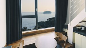A room with a large window offering a view of the sea and a distant island. There is a round table with two chairs and a staircase to the right. - Poong Gyung Hotel (Seogwipo)