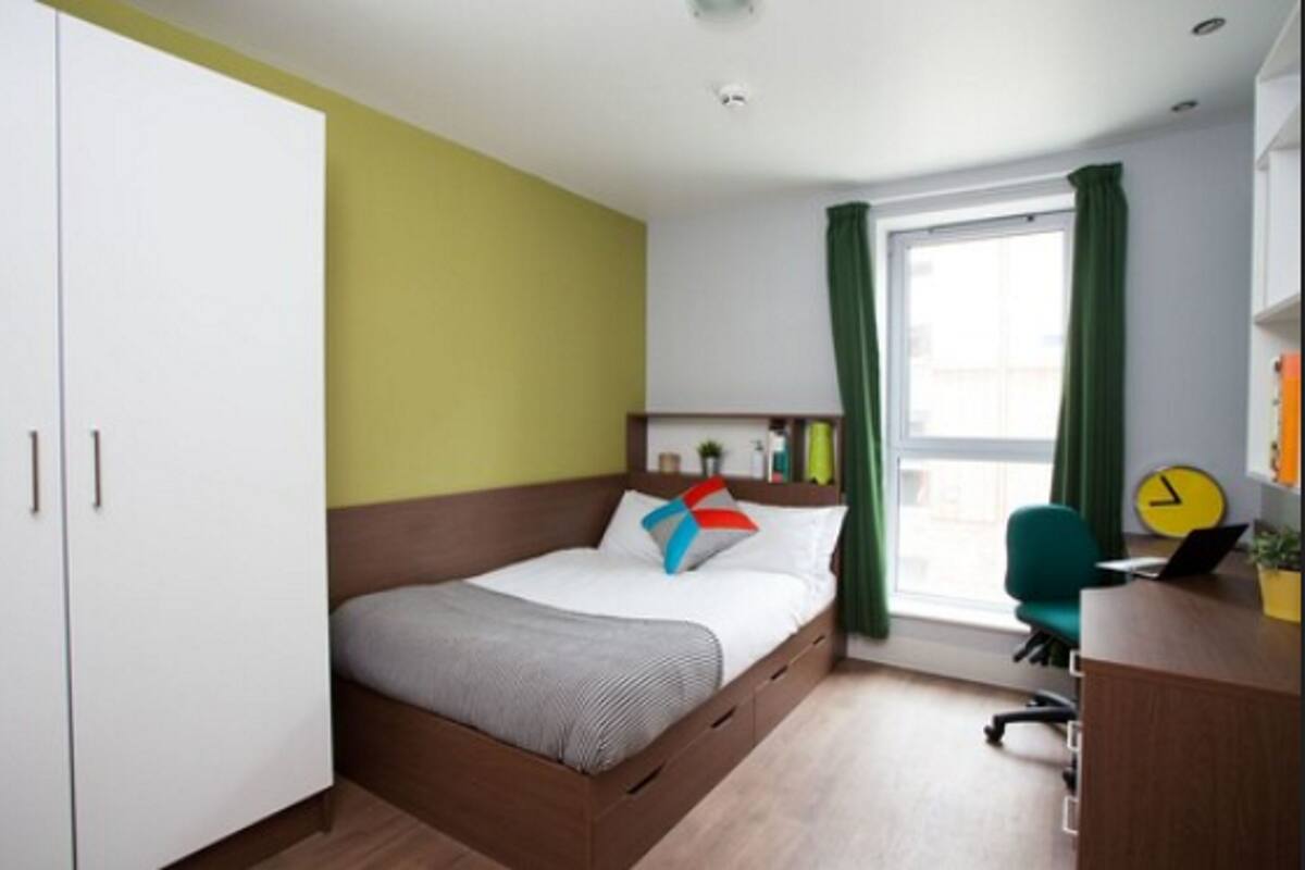 A bedroom with a bed, a desk with a chair, a window with curtains, and a clock on the wall.