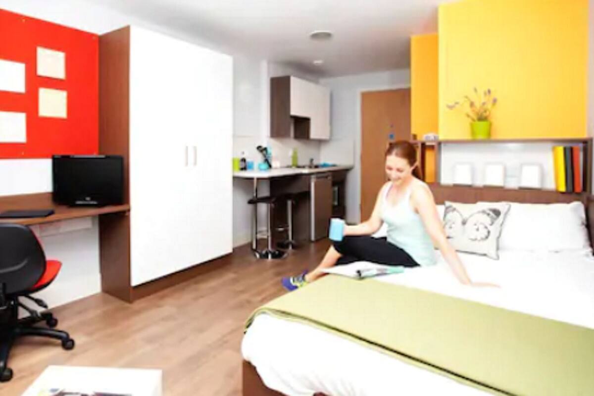 A modern hotel room with a bed, desk, and chair. There is a kitchenette in the background.