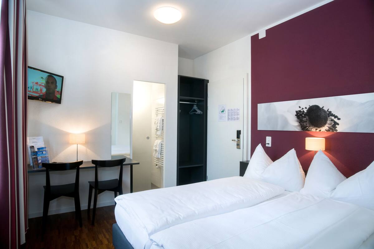 Una camera d'albergo con un letto, una scrivania, due sedie, un armadio e una TV appesa al muro.