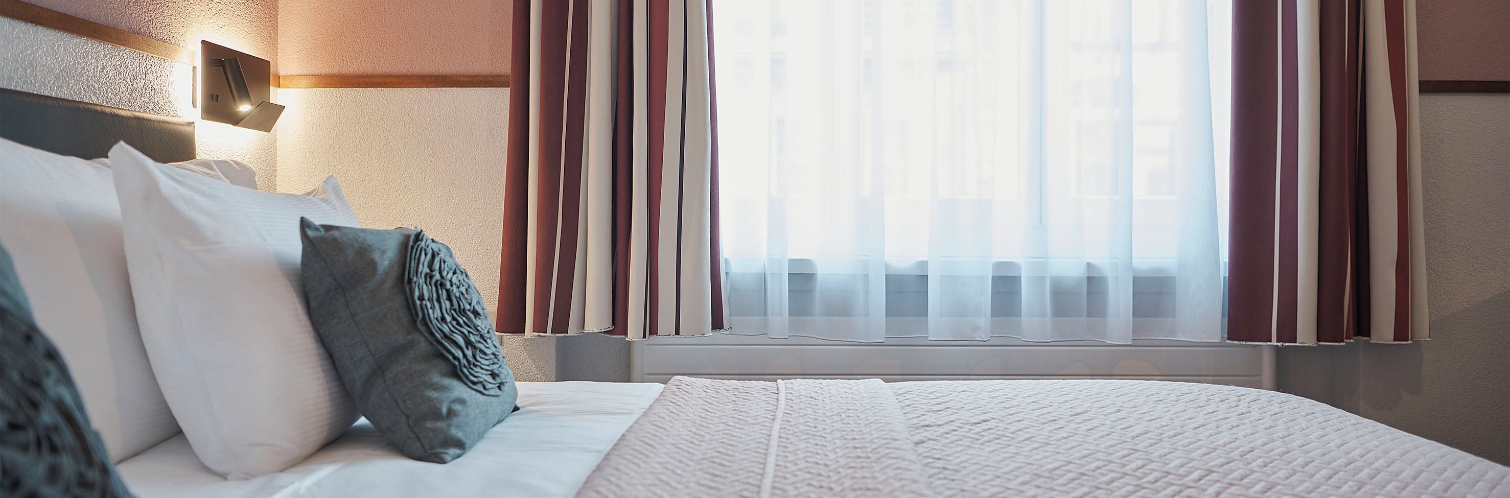A hotel room with a neatly made bed, a headboard with a light fixture, and sheer curtains over a window.