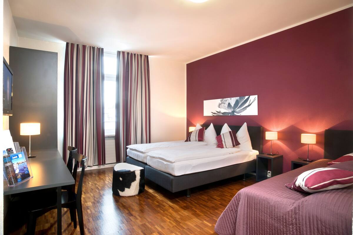 Una camera d'albergo con un letto, due comodini con lampade, una scrivania con una sedia, una TV e una finestra con tende.