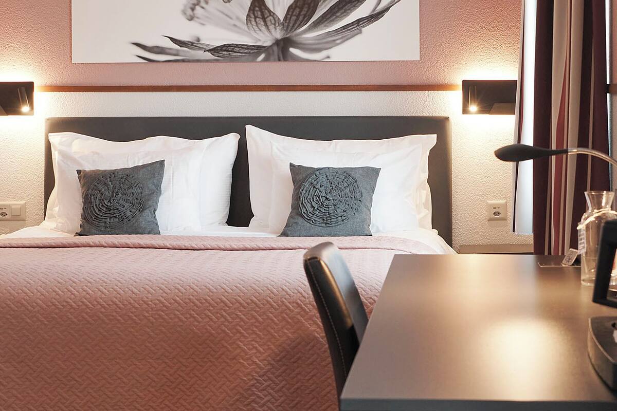 Un letto rifatto con cura, con una coperta rosa, due cuscini e una testiera. Sopra il letto, due luci fissate al muro e un quadro incorniciato appeso al muro.