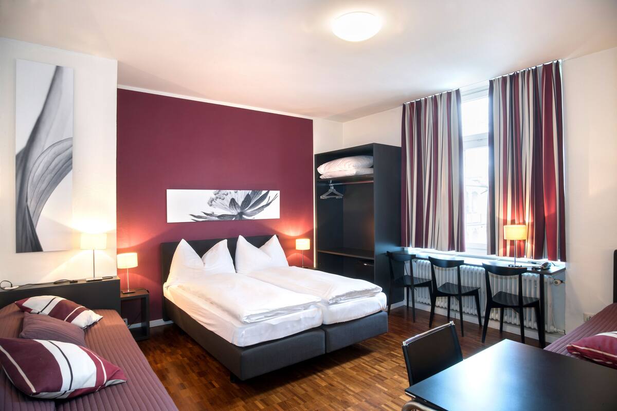 Camera d'albergo con un letto, comodini, una scrivania, sedie, un termosifone e una finestra con tende.