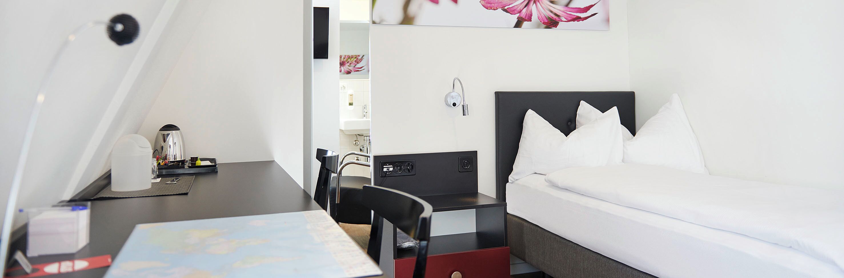 A hotel room with a bed, a desk with a chair, a small table with a map, a nightstand, and a wall-mounted lamp.