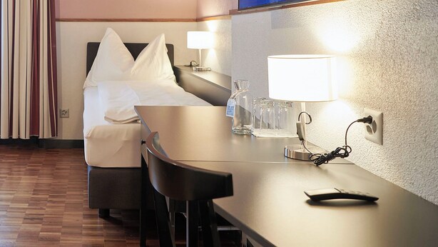 Habitación de hotel con cama, escritorio y una silla. Se observa un radiador, cortinas y un televisor en la pared.