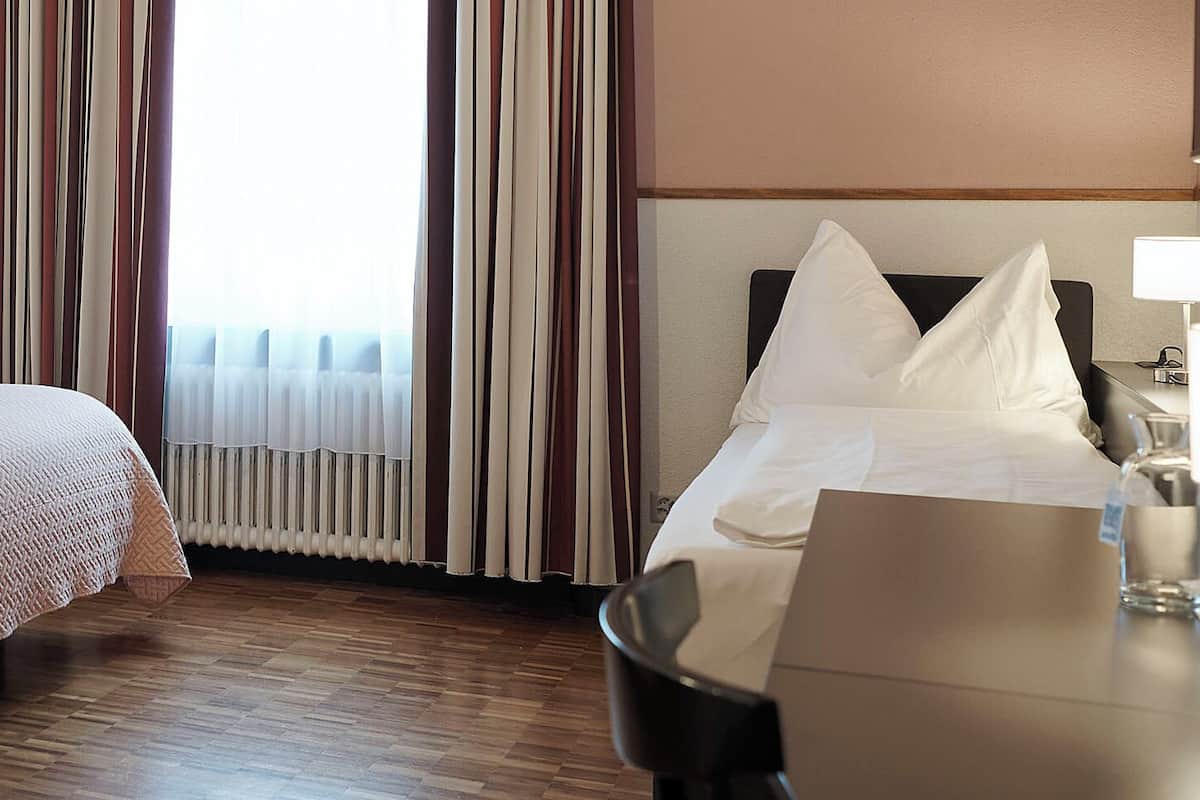 Una camera d'albergo con un letto, un comodino, una lampada e vista sull'esterno dalla finestra.