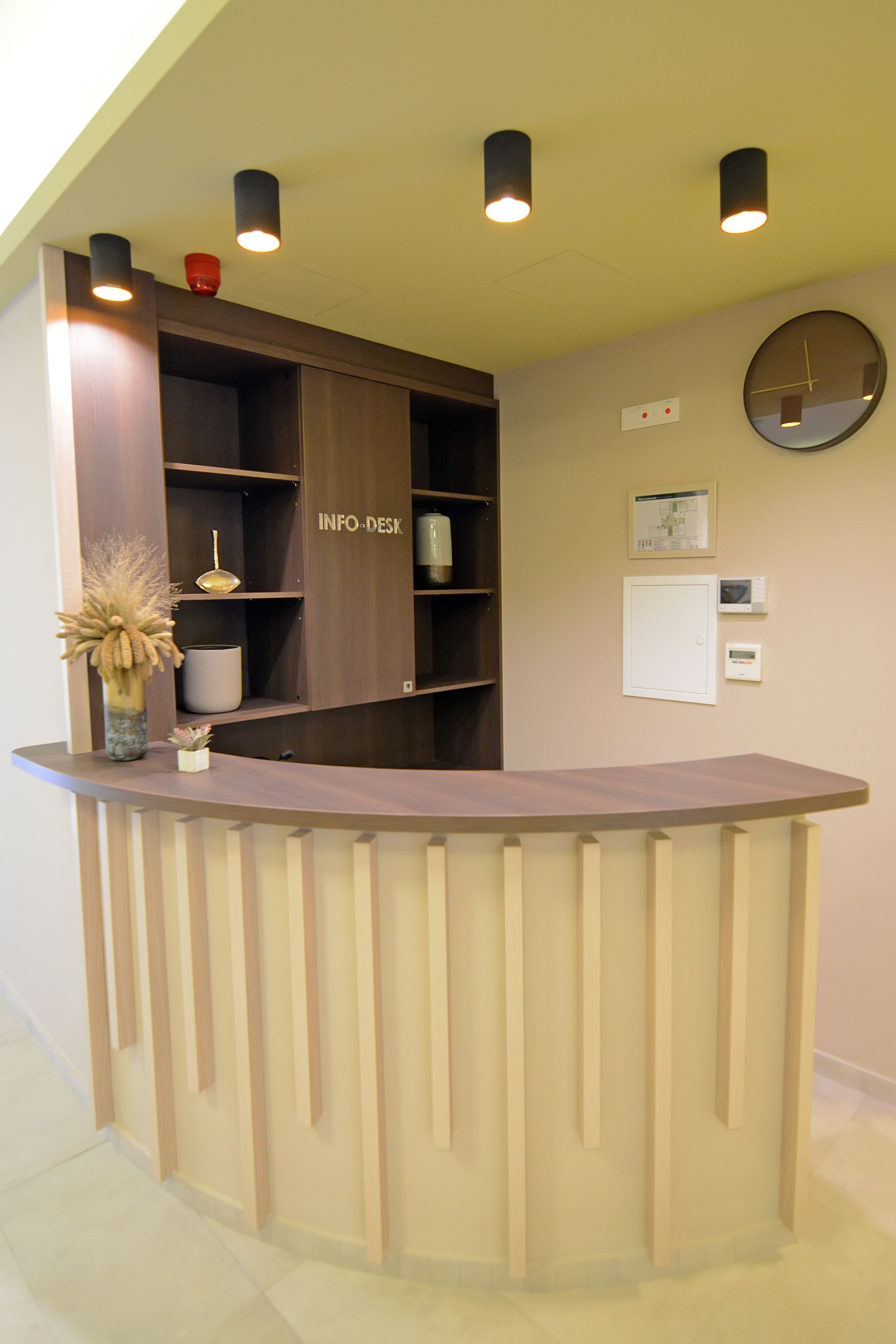 A reception desk with a wooden finish, a vase with dried plants, and a wall-mounted clock.