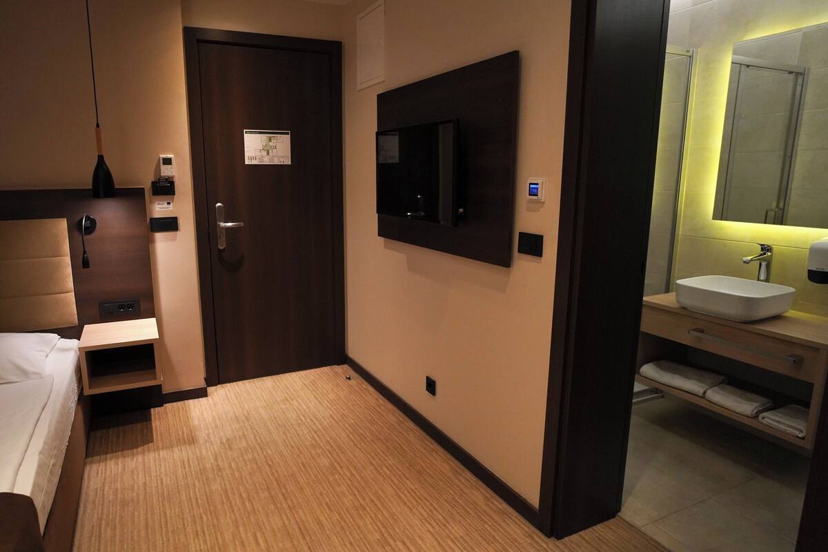 A hotel room with a bed, a bedside table, a TV mounted on the wall, a bathroom with a sink and mirror, and a door with a lock and a keycard reader.