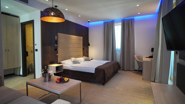 A modern hotel room with a large bed, a desk, a chair, a television, and a dining table with fruit. - Hotel Gallus (Zagreb)