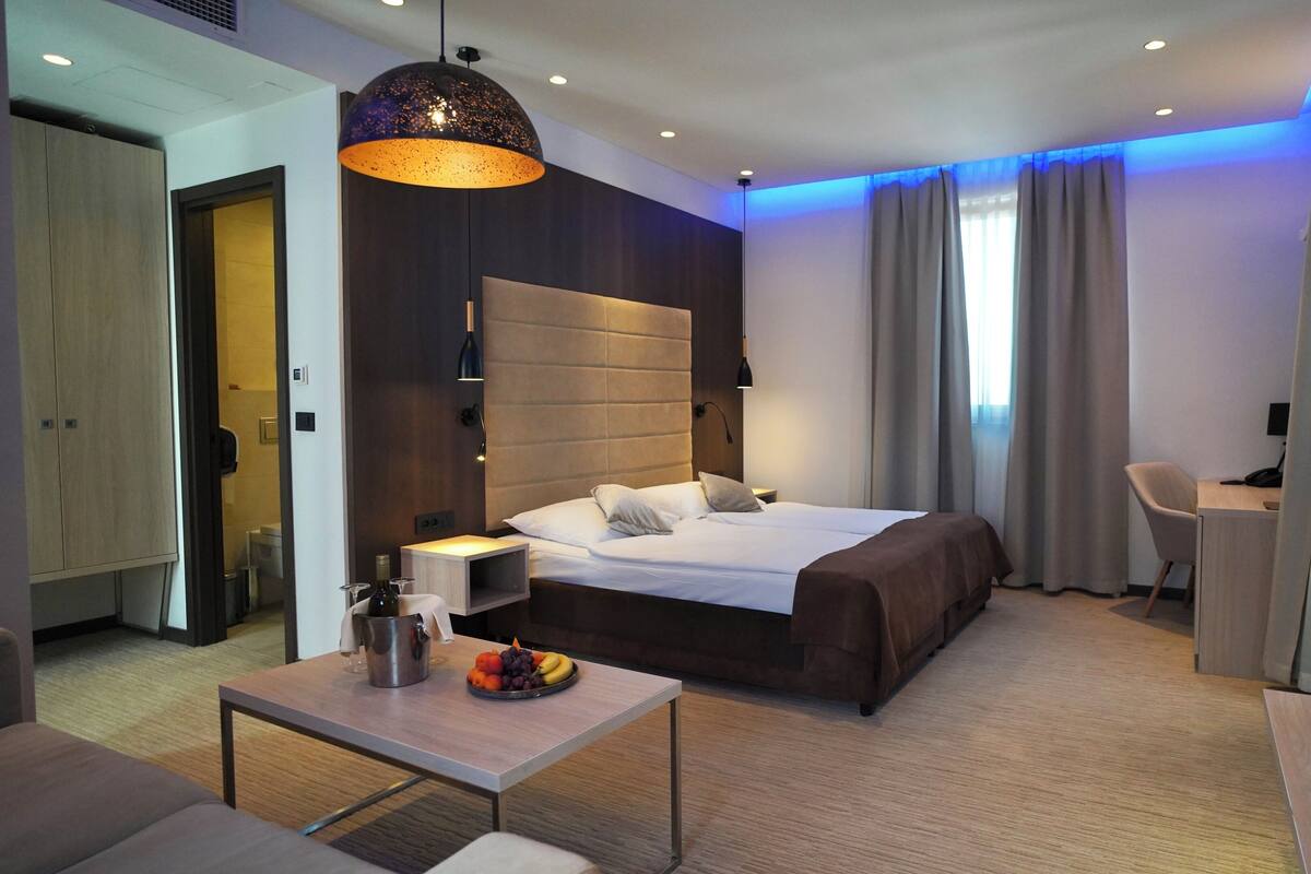 A modern hotel room with a large bed, a desk, a chair, a television, and a dining table with fruit.