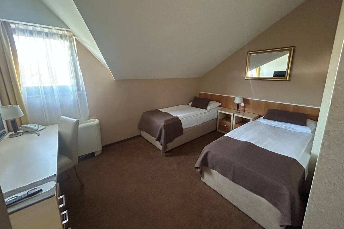 A small, triangular room with two single beds, a desk, a chair, and a bedside table with a lamp.