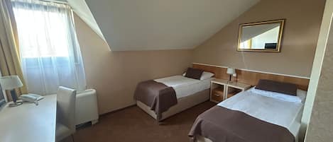 A small, triangular room with two single beds, a desk, a chair, and a bedside table with a lamp.