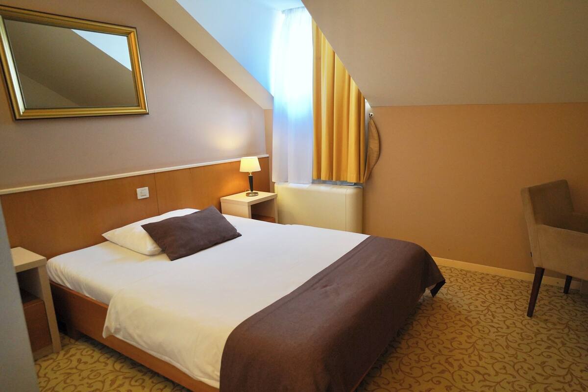 A hotel room with a bed, a desk, a chair, a lamp, and a mirror.