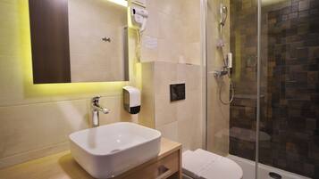 Een badkamer met een witte wastafel, een spiegel, een handföhn, een douche met glazen wanden en een toilet.