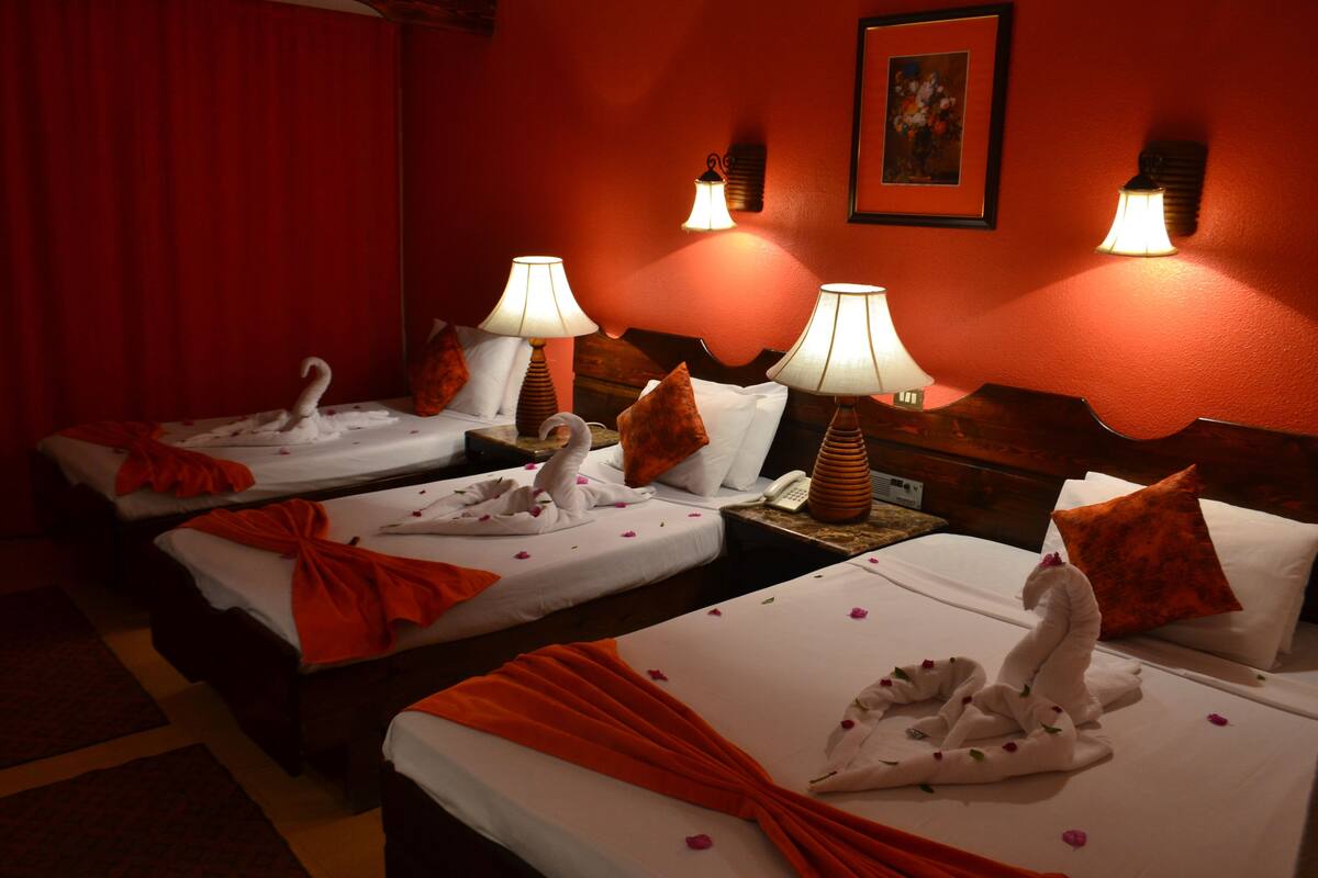A hotel room with three beds, each with a swan towel and orange drapes. There are wall-mounted lamps and a painting on the wall.